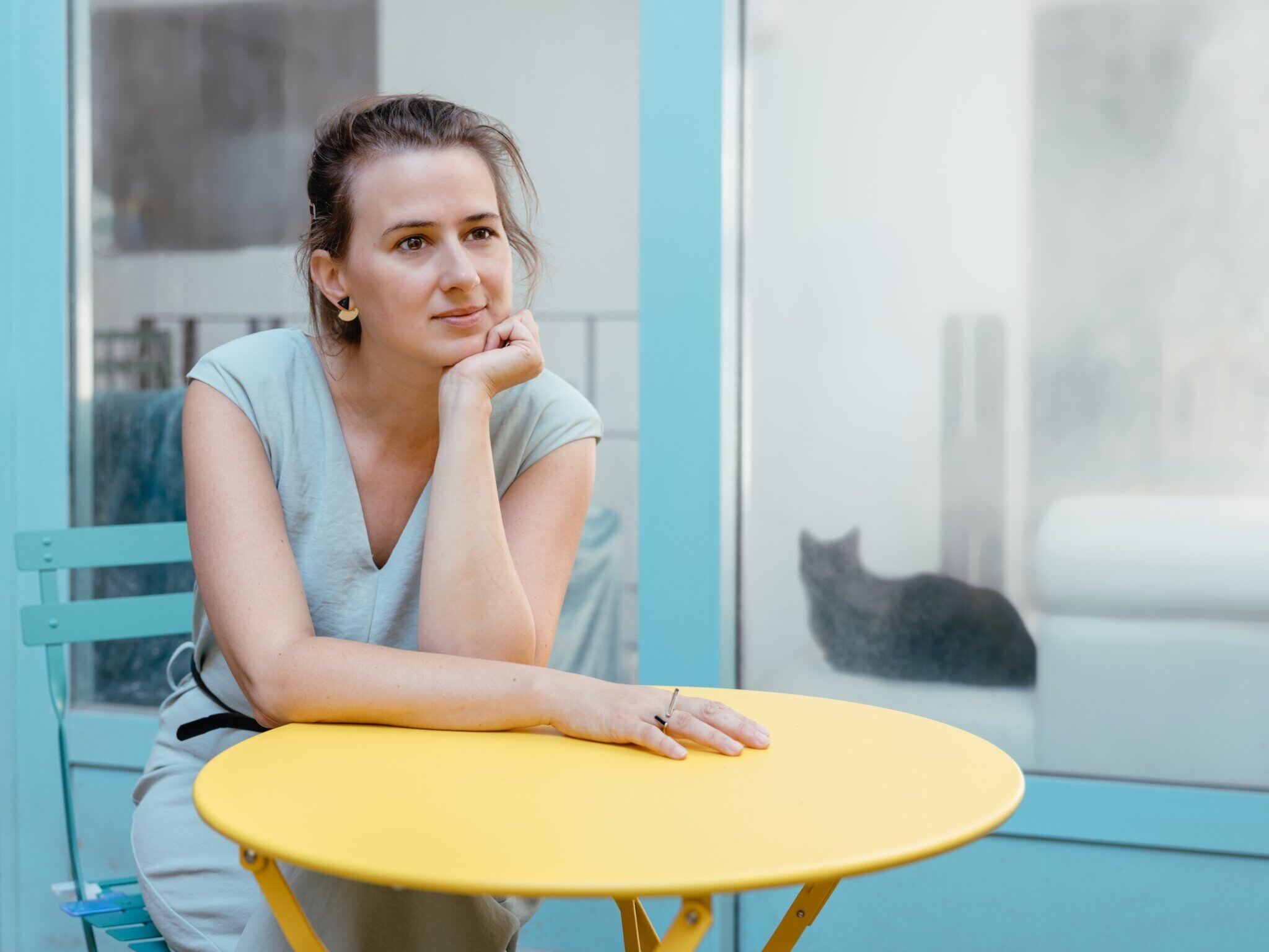 Maud Vanhauwaert. Maud Vanhauwaert dromerig aan een terrastafeltje. Kat op de vensterbank.