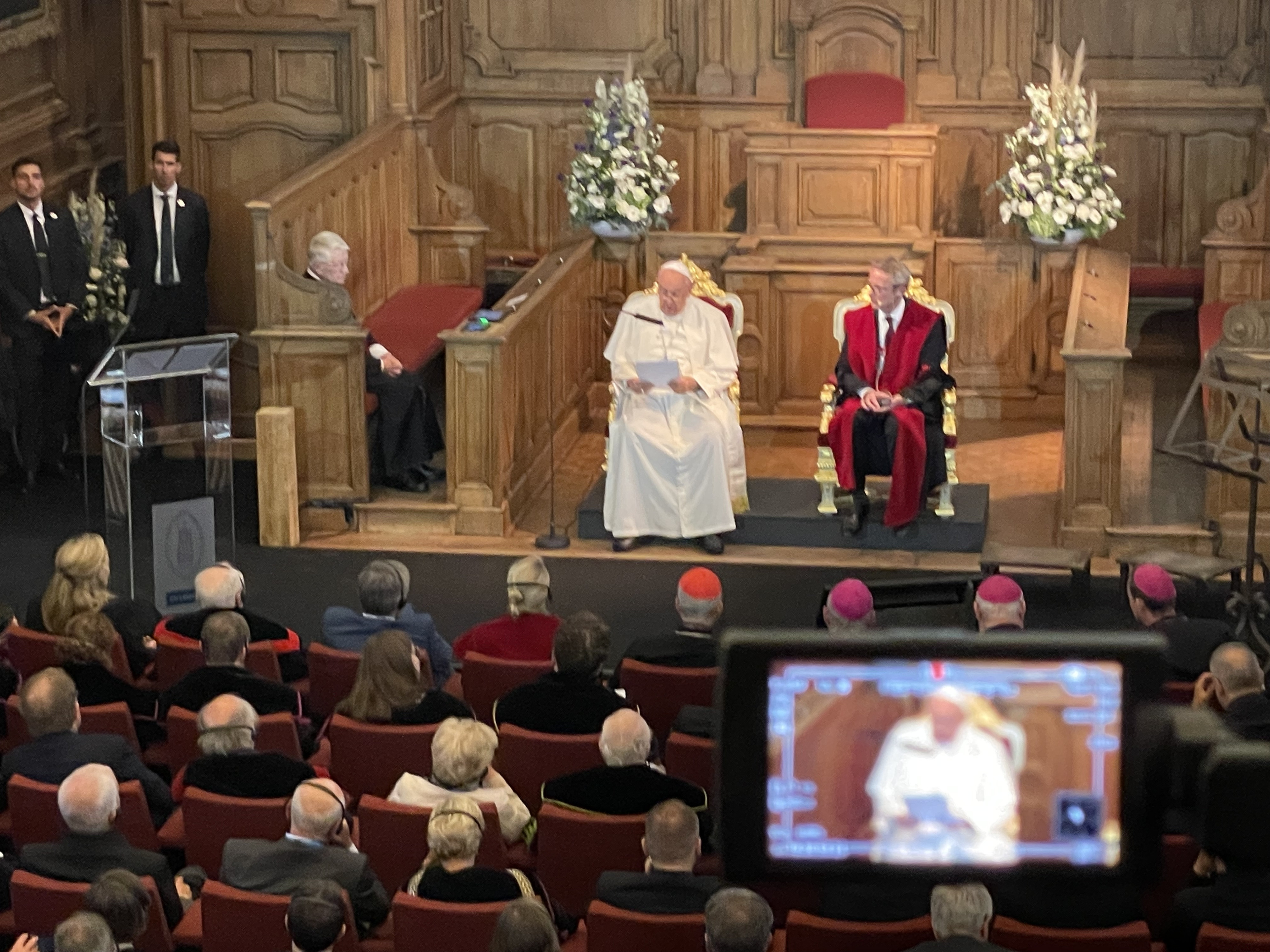 Paus Franciscus aan het woord tijdens de academische zitting in de Promotiezaal van de KU Leuven. Paus Franciscus en rector Luc Sels zitten vooraan in de Promotiezaal van de KU Leuven. Op de voorgrond een camera met hetzelfde beeld.