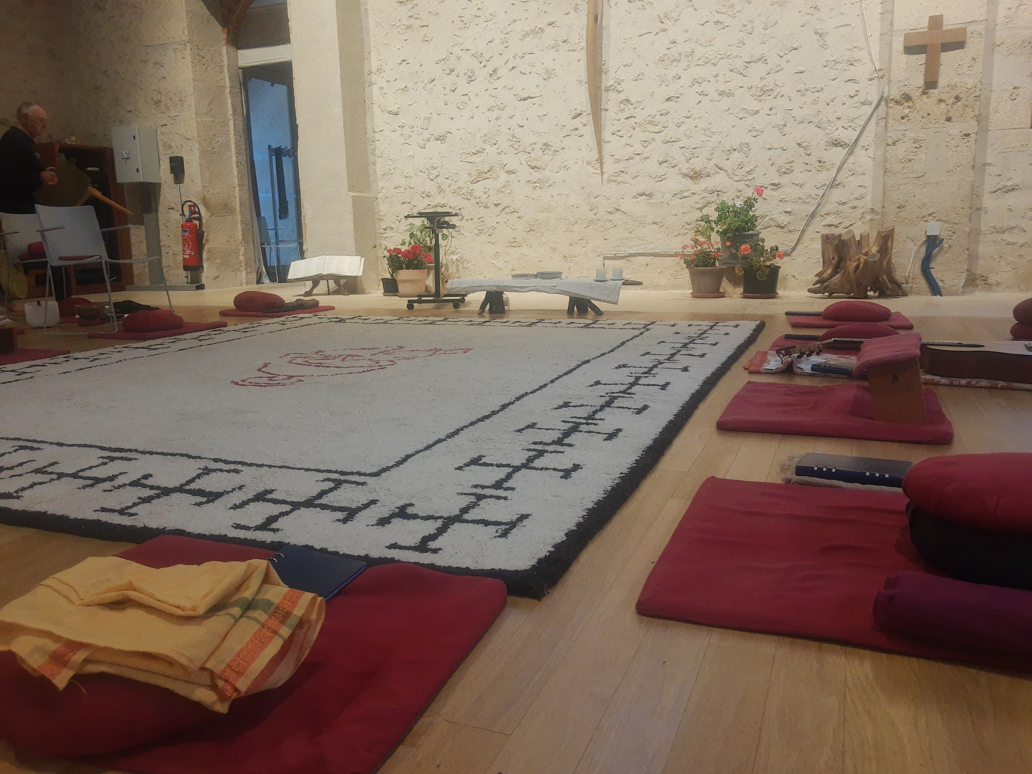 Op de grond ligt een groot rechthoekig tapijt waarop het logo van de christelijke meditatiegemeenschap in het rood geweven is. Abdij Bonnevaux_meditatietapijt
