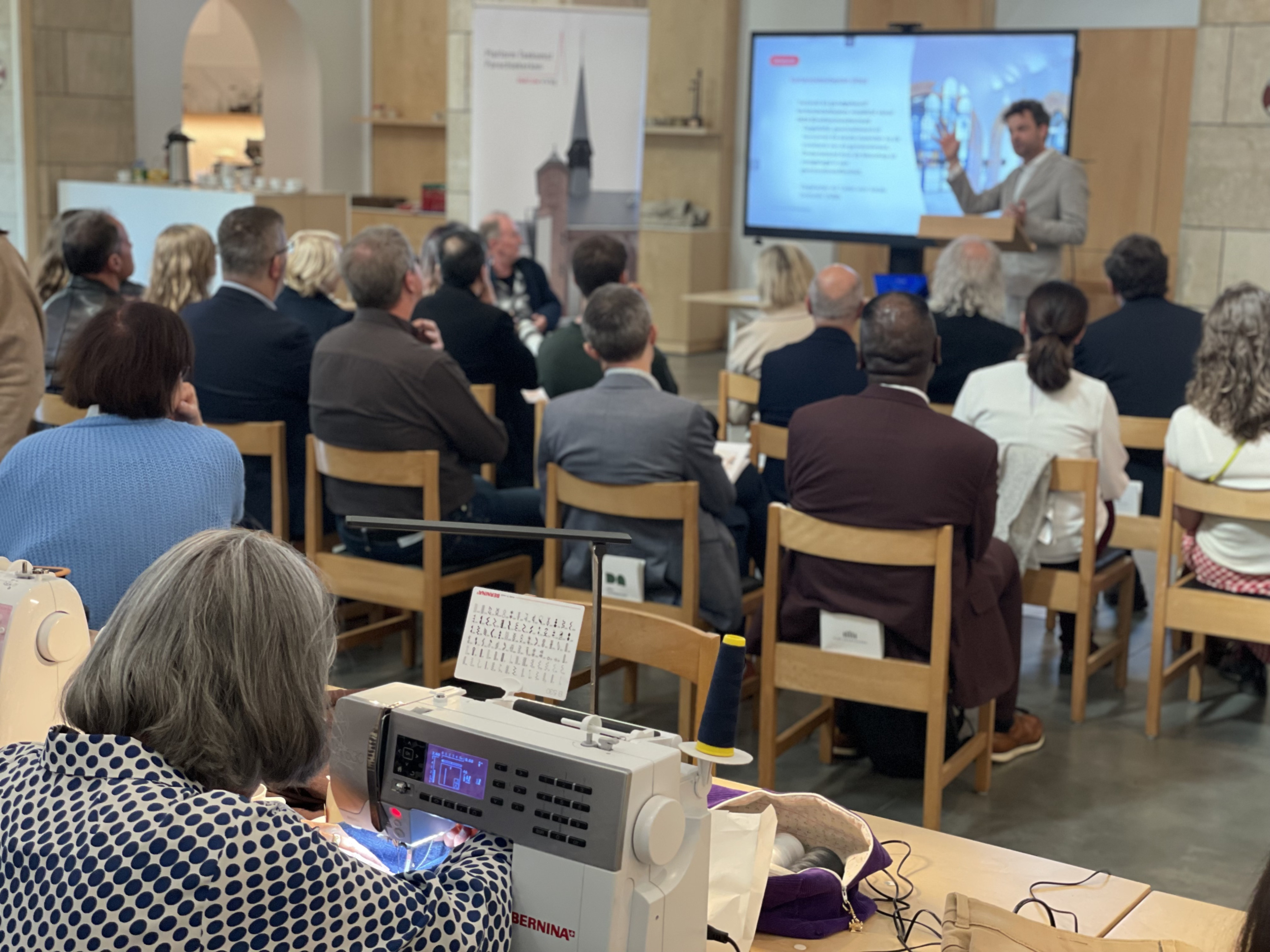 Op de voorgrond is een vrijwilliger van het Repair Café bezig aan een naaimachine. Op de achtergrond is de persconferentie bezig. 