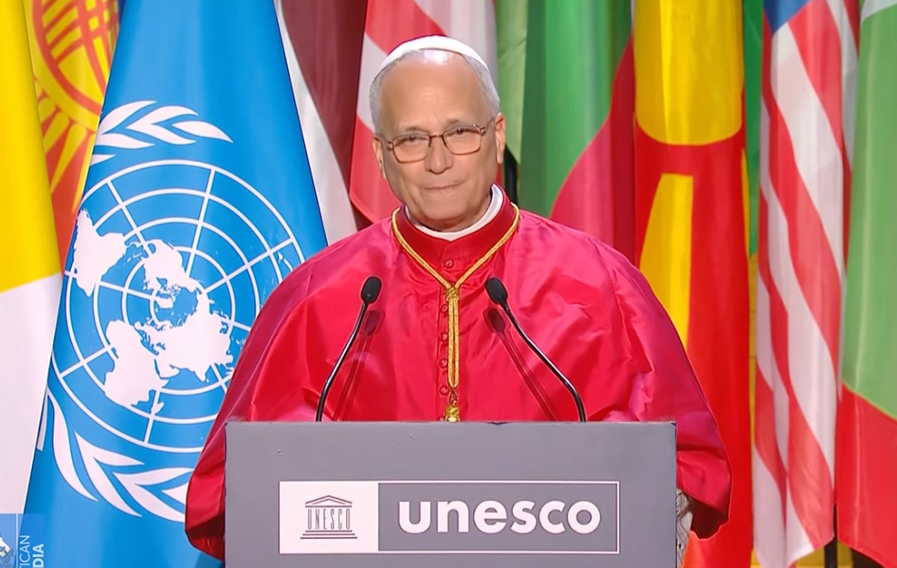 Paus Leo aan de micro bij de UNESCO. Op de achtergrond kleurige vlaggen van alle lidstaten.