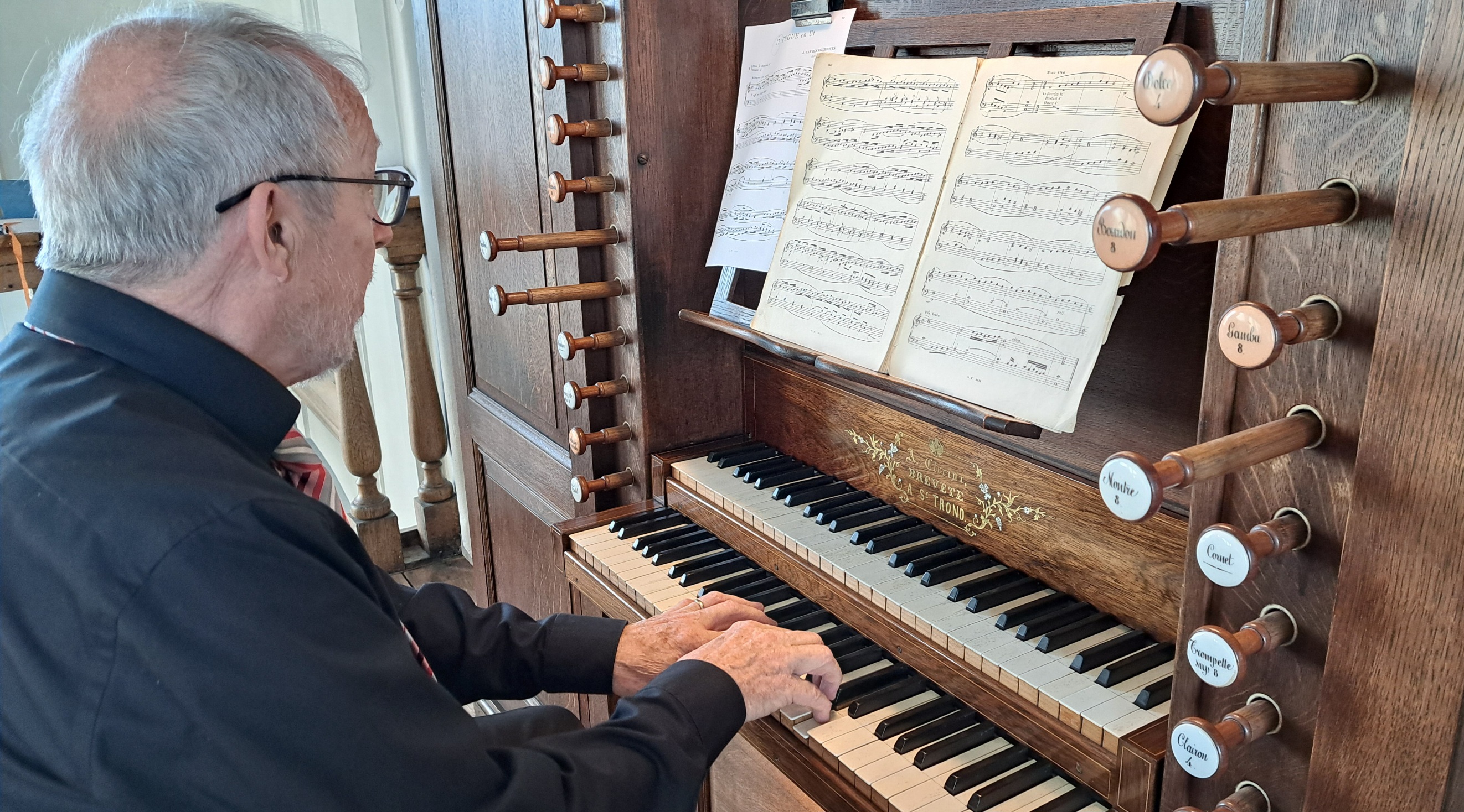 Hugo Adams die aan de speeltafel van een pijporgel zit en op twee manualen (klavieren) speelt, te midden van bladmuziek en herkenbare registertrekkers aan de zijkant.