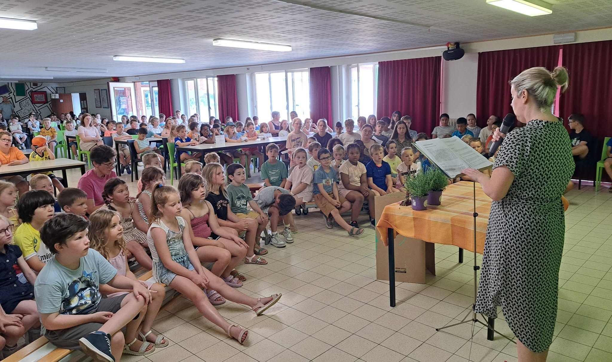Een klaslokaal waarin een groep basisschoolleerlingen op de grond en op banken zit te luisteren naar een leerkracht of spreker die achter een tafel staat.