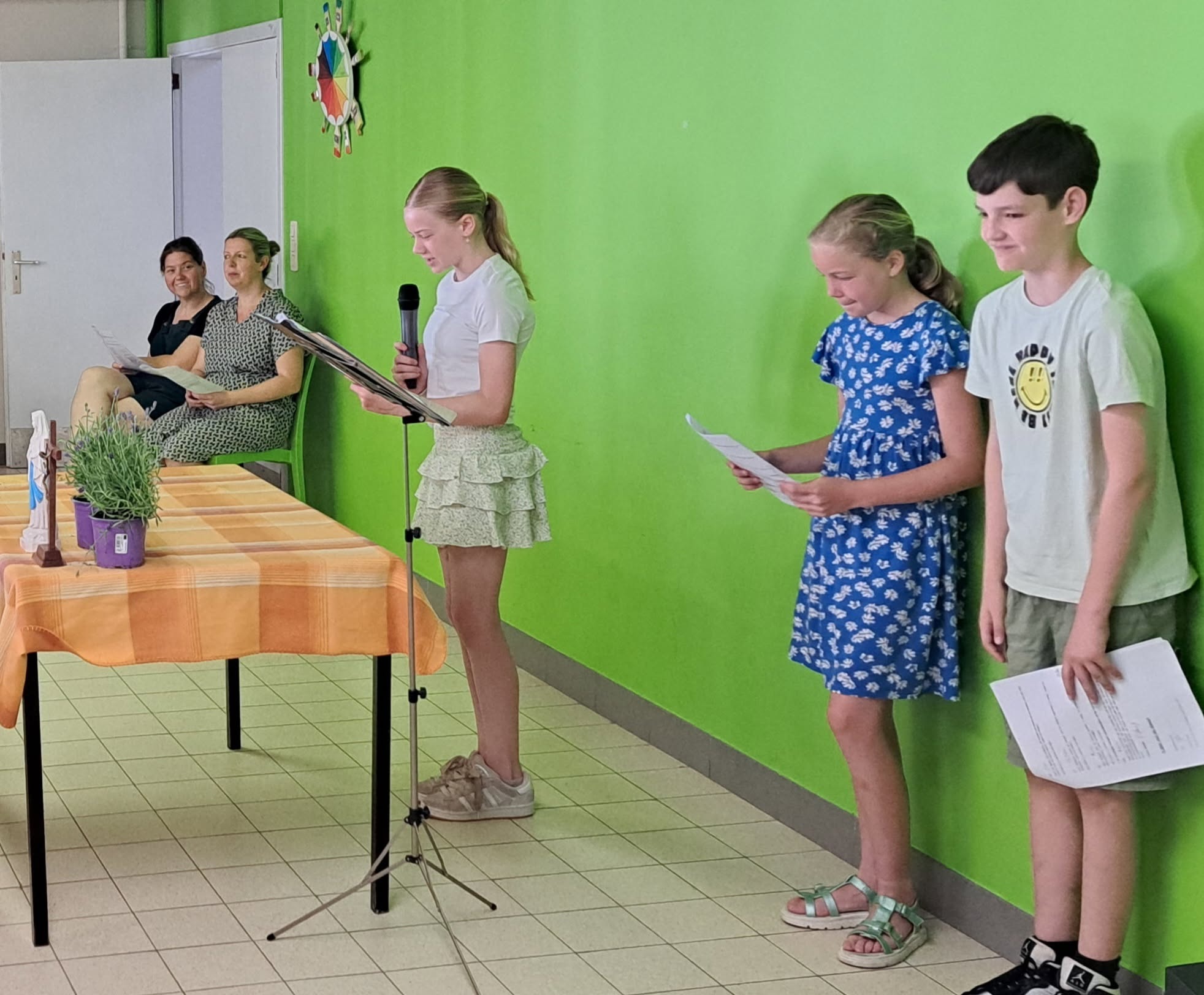 Een schoolactiviteit of toonmoment in een klaslokaal waar kinderen met papieren in hun hand bij een microfoon staan en toeschouwers toekijken.