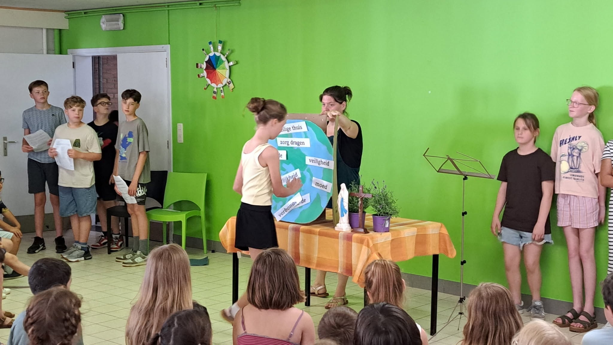 Een klaslokaal of ruimte met groene muren waarin een groep kinderen en een volwassen begeleider of leerkracht rond een tafel staan opgesteld voor een gebedsviering.