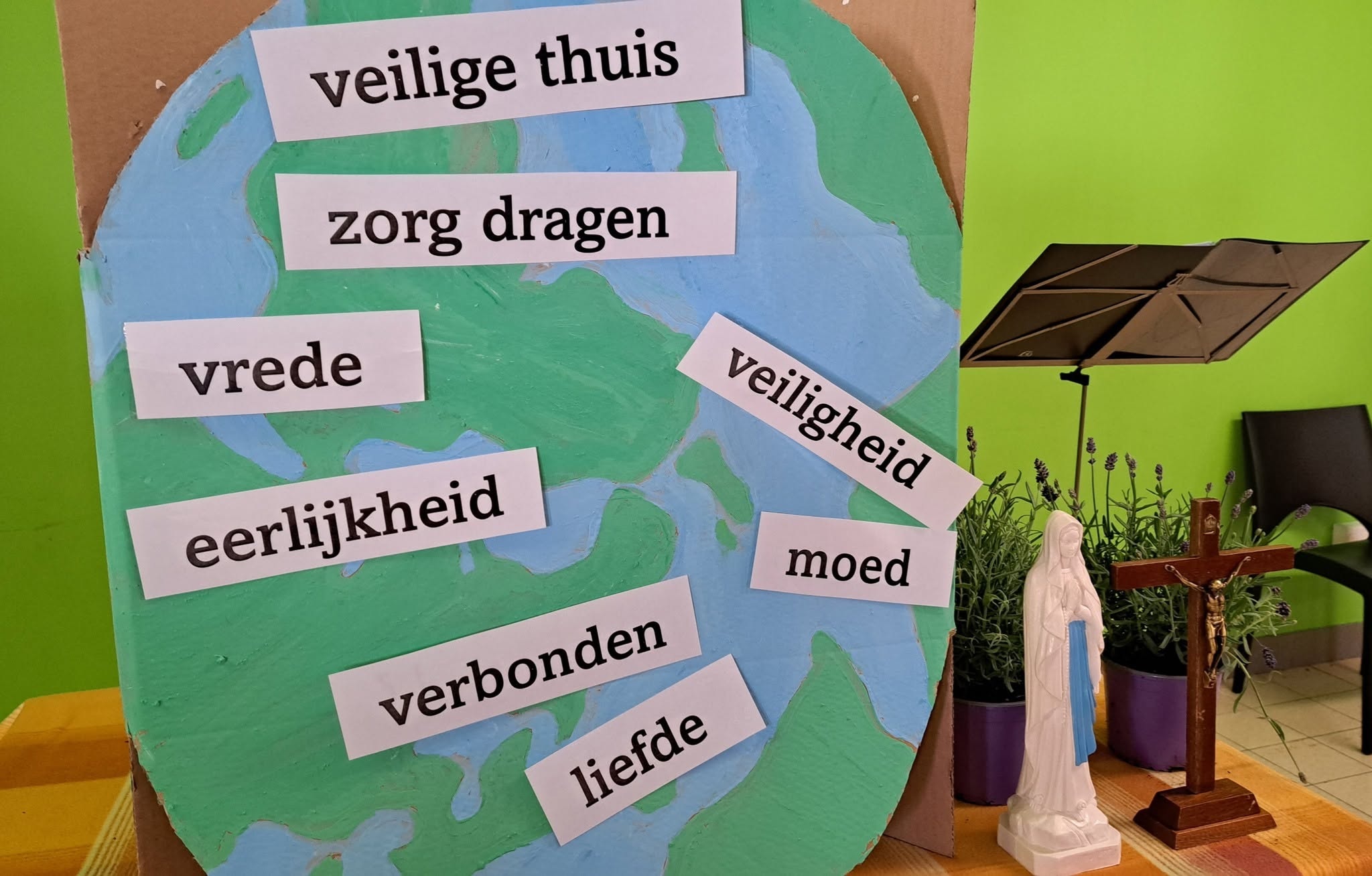 Een knutselwerk of bezinningshoek met een wereldbol waarop waarden staan zoals "veilige thuis", "zorg dragen", "vrede", "eerlijkheid", "verbonden", "liefde", "moed" en "veiligheid", naast een Mariabeeld en een kruisbeeld.