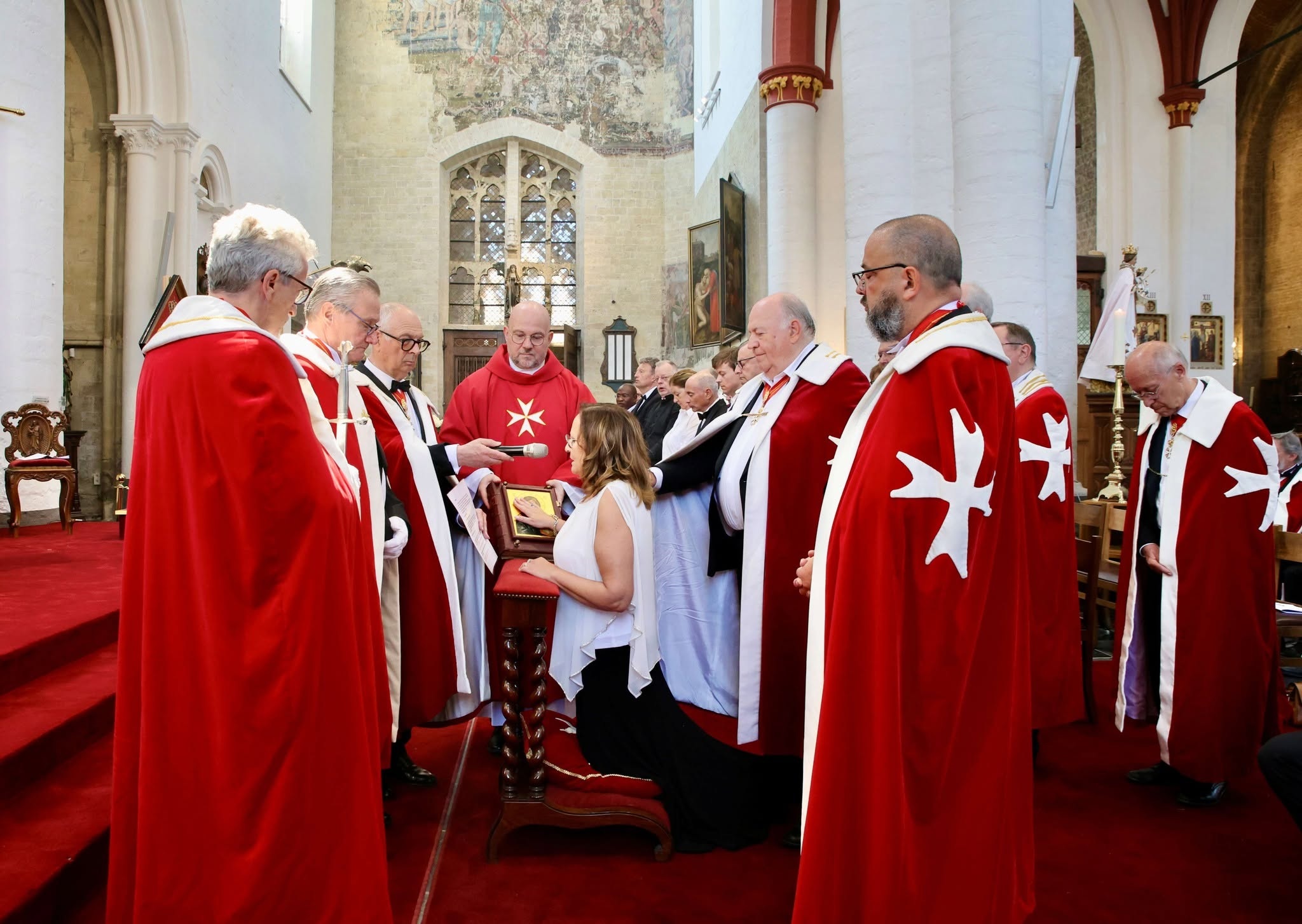 Een investituur- of aannemingsceremonie van een ridderorde — zoals de Hospitaalridders van Sint-Jan van Jeruzalem (of een verwante ridderlijke/oecumenische orde) — waarbij personen in rode mantels met een wit achtpuntig kruis (Maltezer kruis) aanwezig zijn.