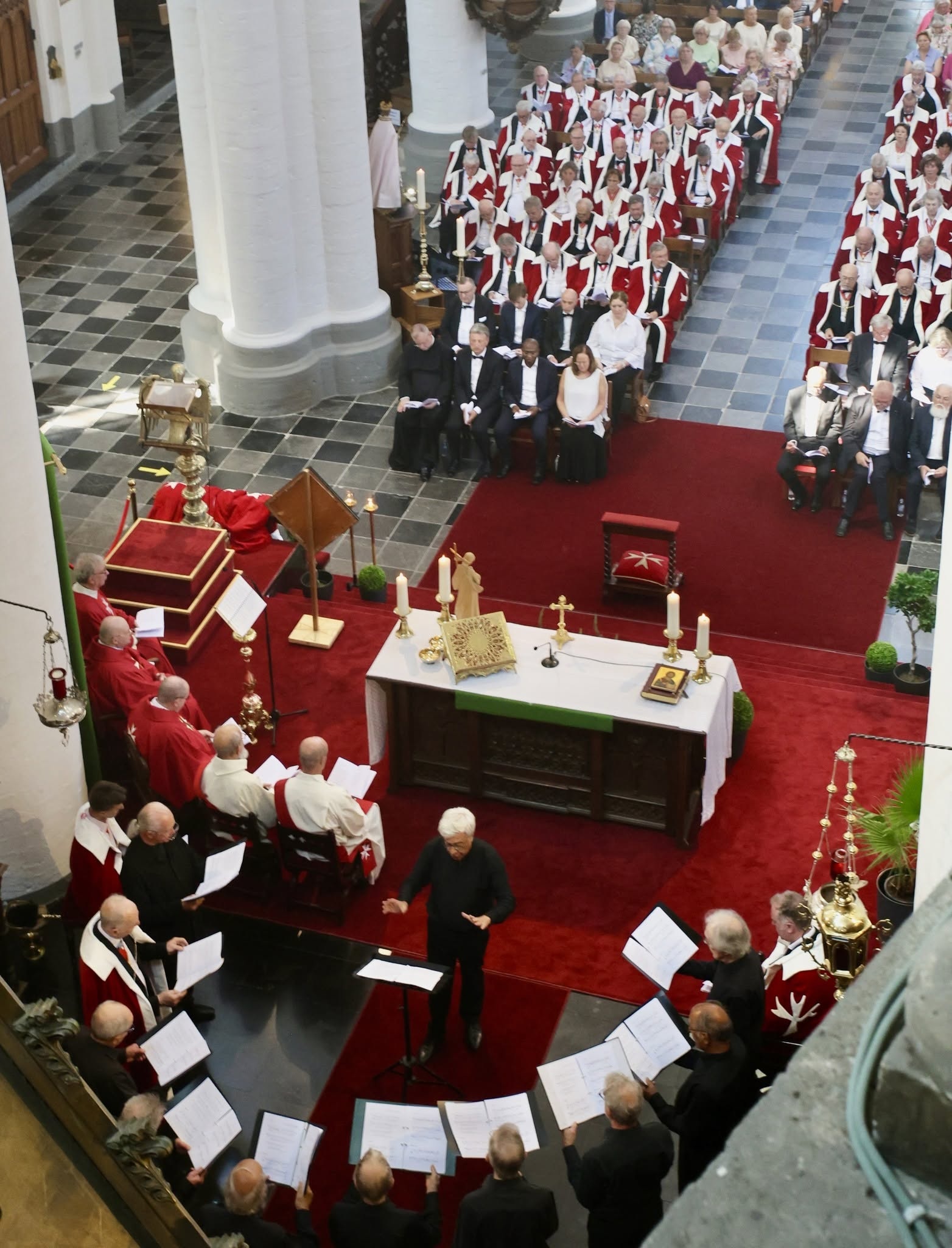 Een plechtige kerkdienst te tonen — vermoedelijk een investituur, herdenking of plechtige viering van de Orde van Sint-Jan van Jeruzalem (Hospitaalridders - OSJ) — met een koor dat onder leiding van een dirigent zingt bij het altaar en rijen genodigden of leden in gewaden.