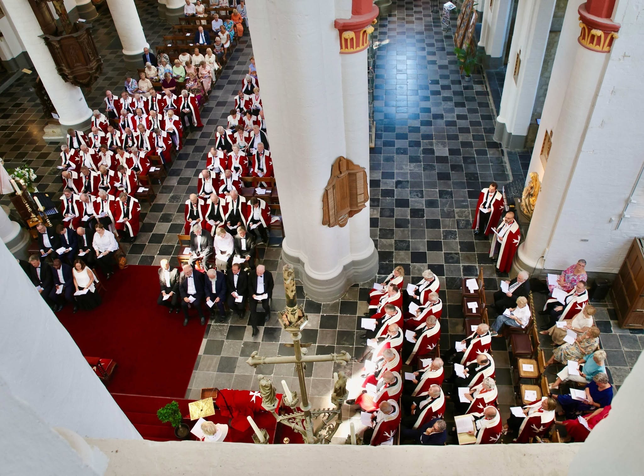 Een plechtige bijeenkomst, waarschijnlijk een kerkdienst of investituur (zoals van een ridderorde of academische plechtigheid), met mensen in formele of liturgische kleding die in de kerk zijn opgesteld rondom de zuilen en het schip.