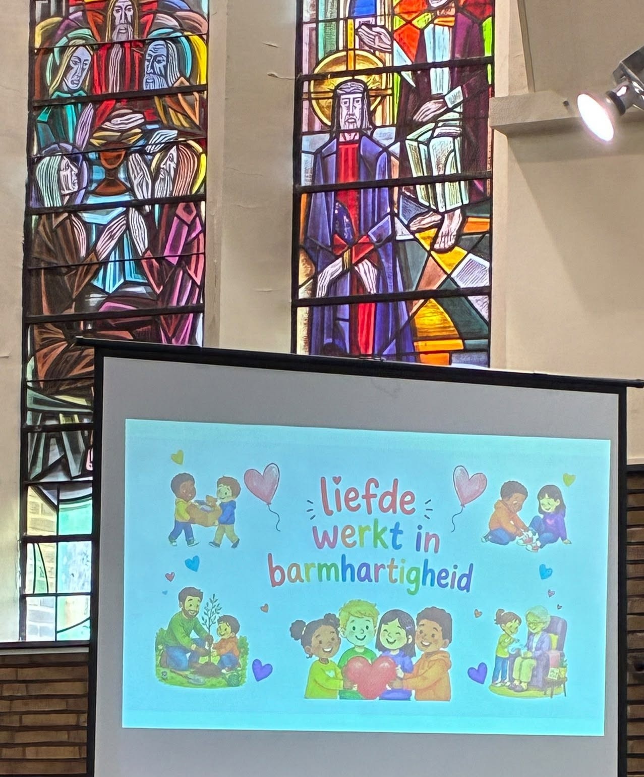 Een projectiescherm in een kerk met de tekst "liefde werkt in barmhartigheid" en illustraties van kinderen en volwassenen die elkaar helpen.