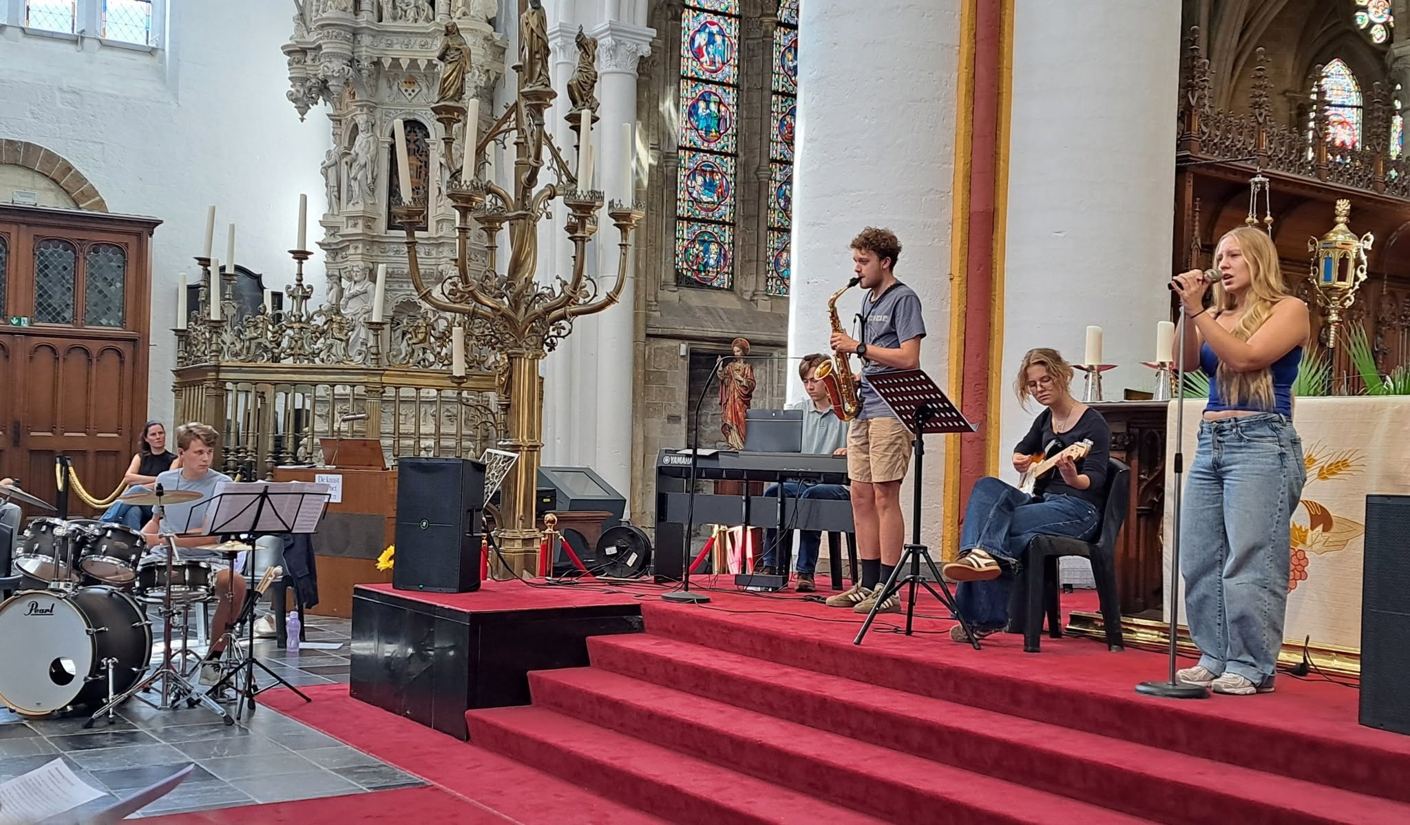 Middelbare scholen Zoutleeuw maken een gelovige start Een muziekgroep die optreedt in de Sint-Leonarduskerk in Zoutleeuw. Op het podium staan enkele jonge muzikanten met instrumenten zoals een gitaar en een saxofoon, en een zangeres bij een microfoon.