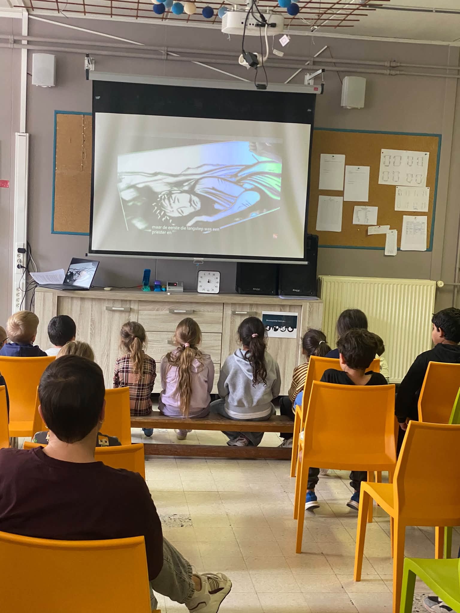 Een klaslokaal of een ruimte met een groep kinderen die op gele stoelen en een bankje naar een projectiescherm kijken.