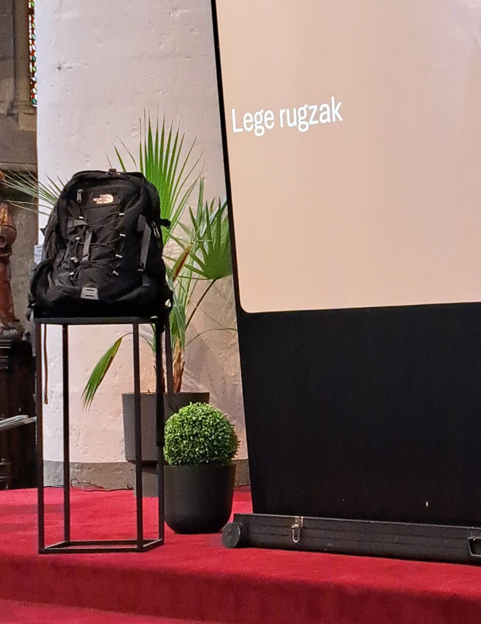 Middelbare scholen Zoutleeuw maken een gelovige start Een zwarte rugzak die op een smalle zwarte zuil staat naast een projectiescherm met de tekst "Lege rugzak" erop.