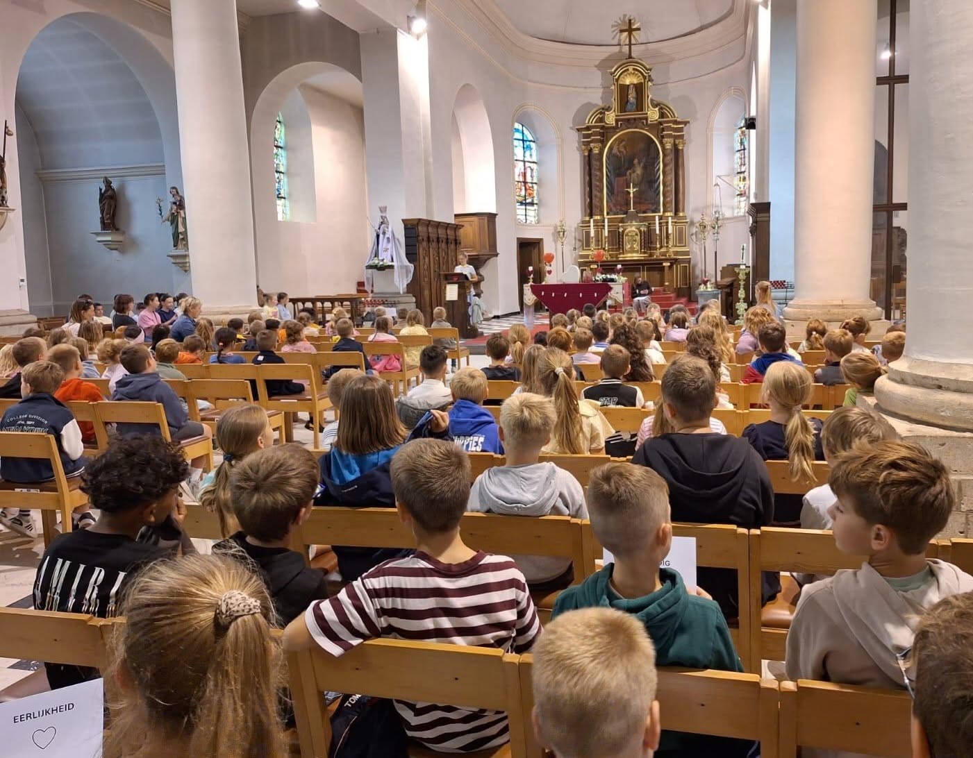 Een groep leerlingen en begeleiders tijdens een schoolviering of kerkdienst in een traditioneel kerkinterieur met houten banken, witte zuilen en een altaar.