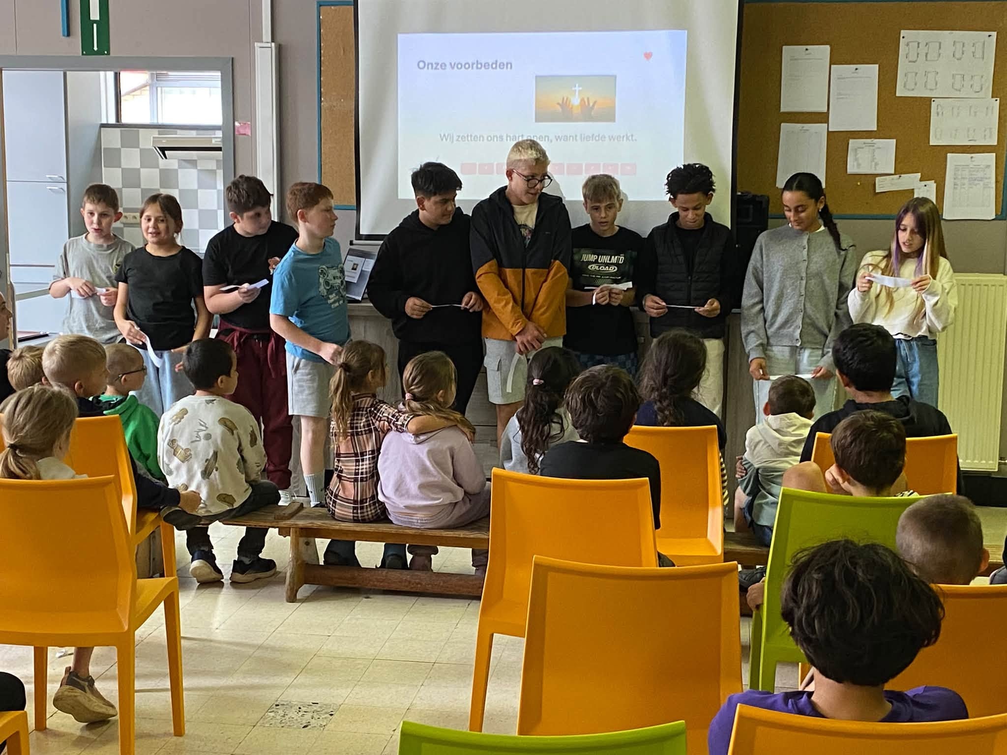 Een klaslokaal waarin een groep leerlingen voor de klas staat, met gele en groene stoelen op de voorgrond en een presentatiescherm op de achtergrond.