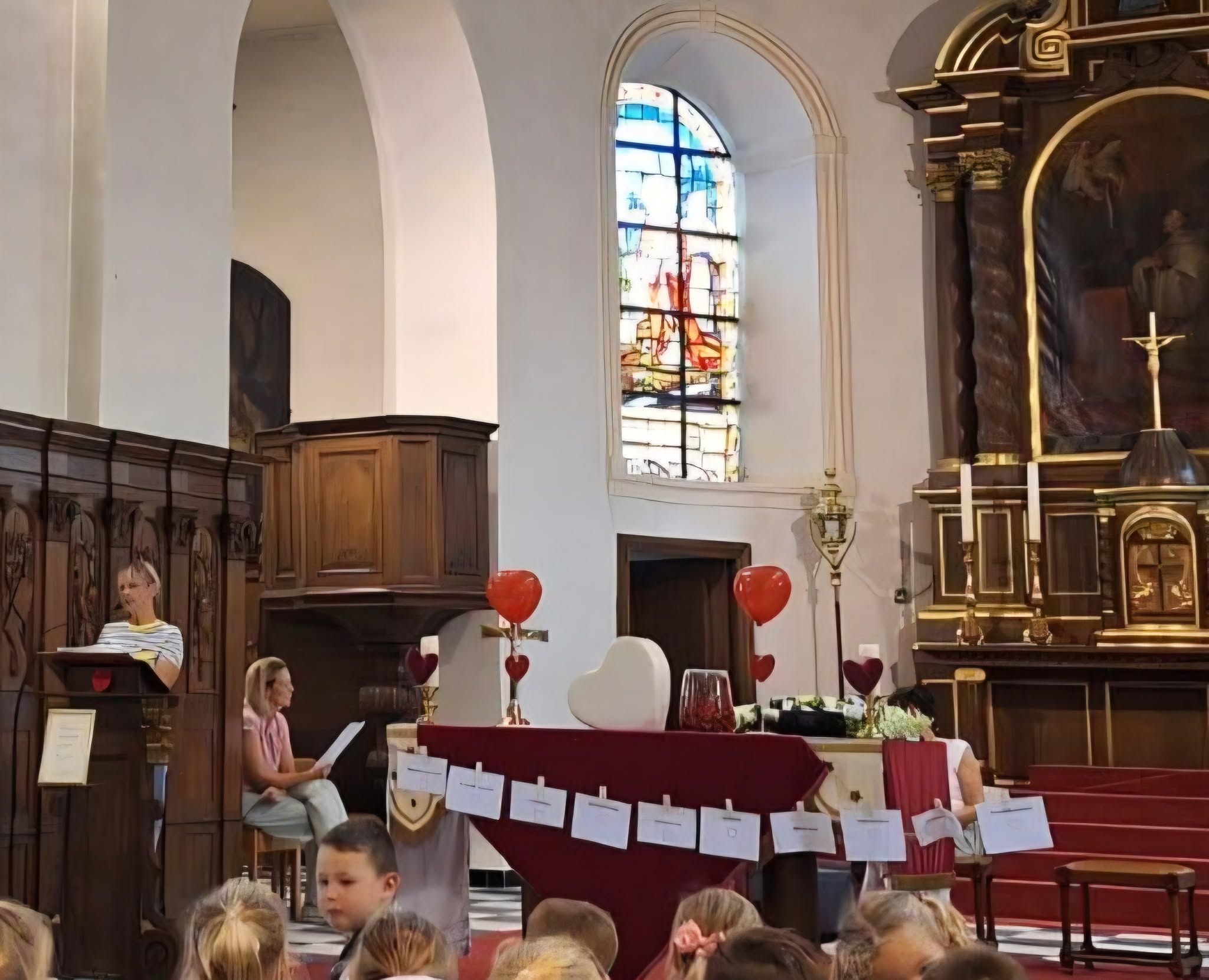 Viering (zoals een school- of communieviering) in een traditionele kerkruimte te tonen, met personen bij een lessenaar en een altaar versierd met rode hartballons.
