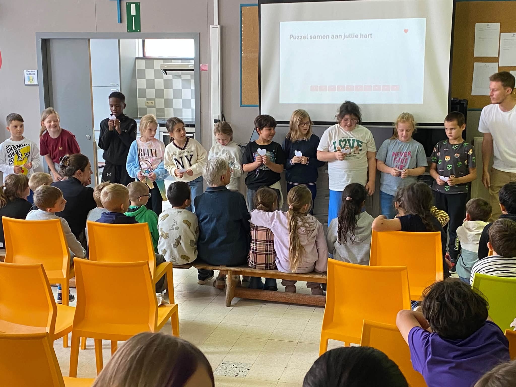 Een groep kinderen en leerkrachten tijdens een schoolbijeenkomst in een klas- of polyvalente ruimte met gele stoelen en een projectiescherm.
