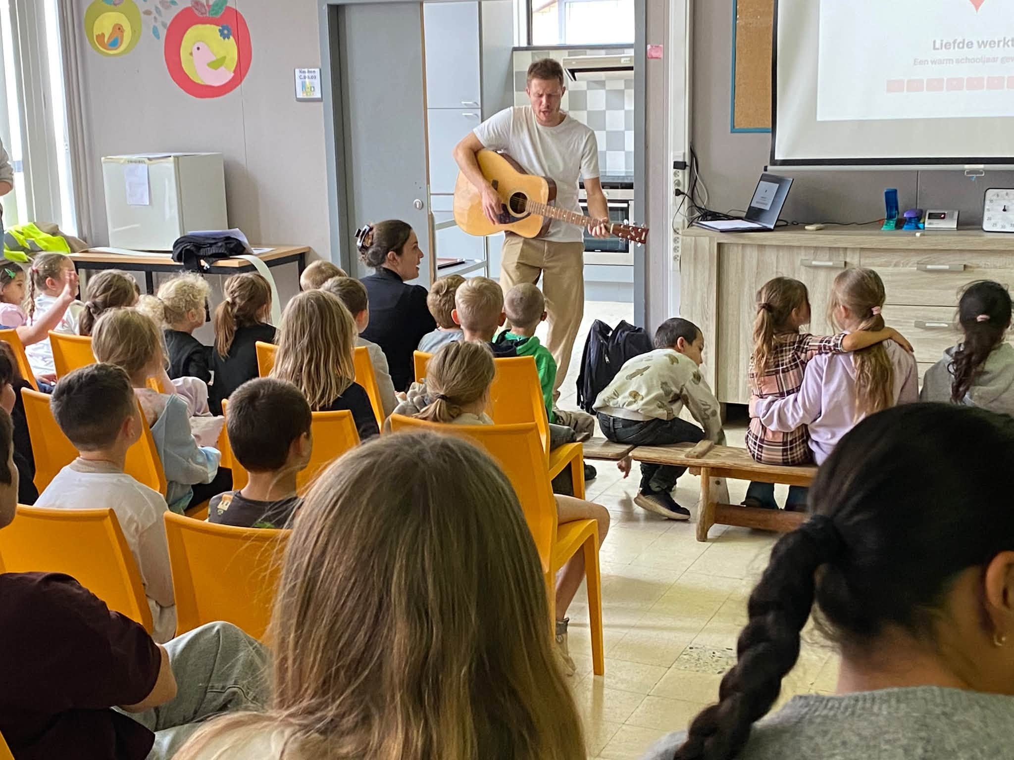 Een klaslokaal of zaal waar kinderen op gele stoelen naar een persoon kijken die met een akoestische gitaar voor hen staat.