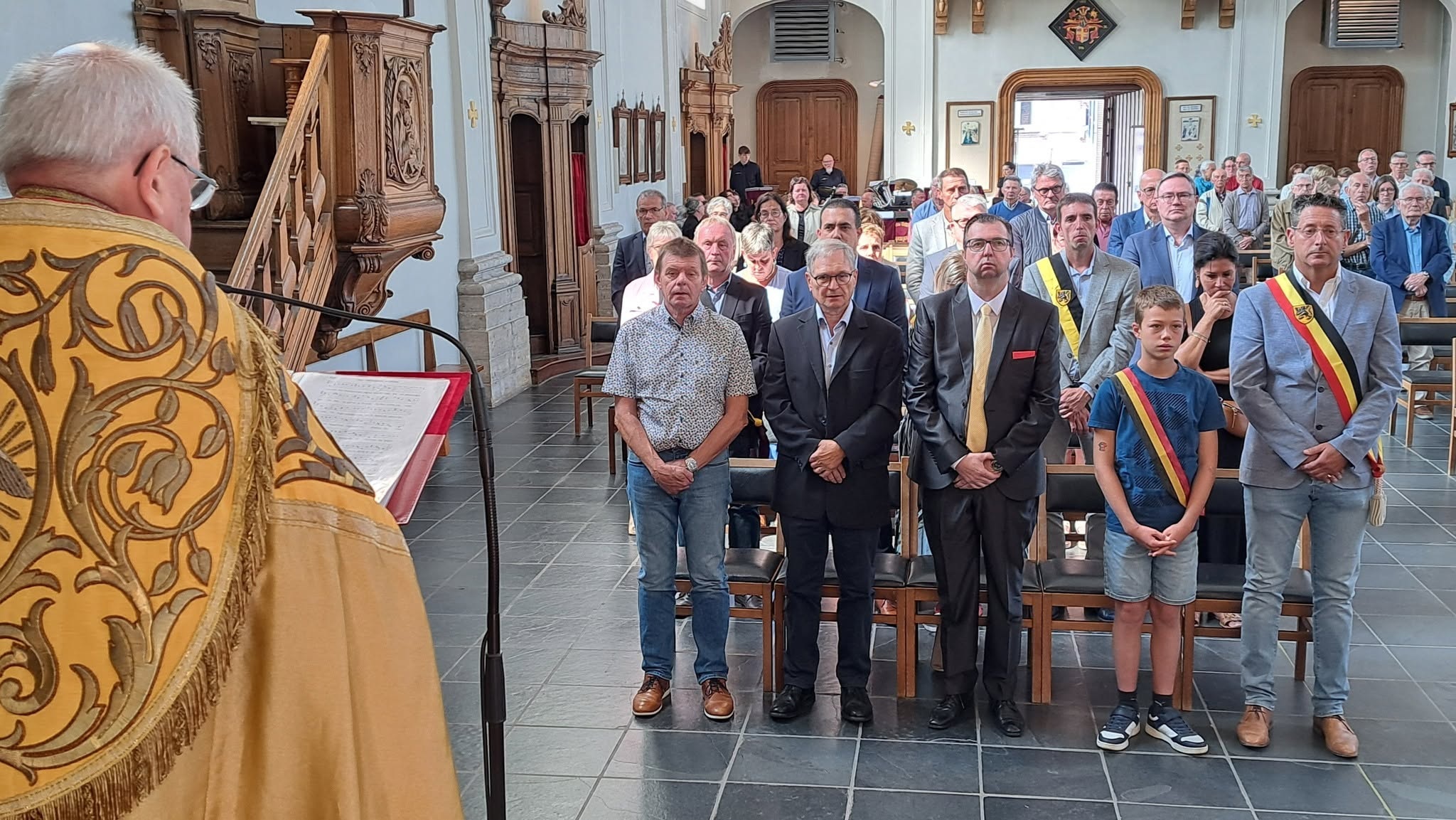 Te Deum-viering op Nationale Feestdag Een plechtige Te Deum-viering (vaak ter gelegenheid van de Belgische nationale feestdag op 21 juli) in een kerk, waar een priester in liturgische kleding voorgaat en de lokale burgerlijke overheden met driekleurige sjerpen aanwezig zijn.