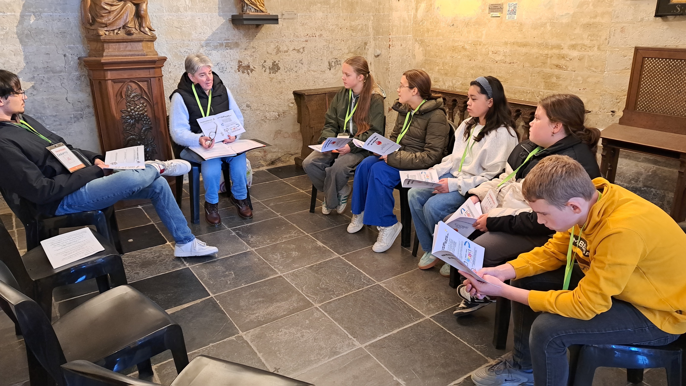 Samen op weg naar het vormsel Een groep mensen, waaronder jongeren en begeleiders of catechisten, die in een kring op stoelen in een ruimte (zoals een kerkruimte of parochiezaal) zitten met papieren of mappen in hun handen tijdens een bijeenkomst of catechese.