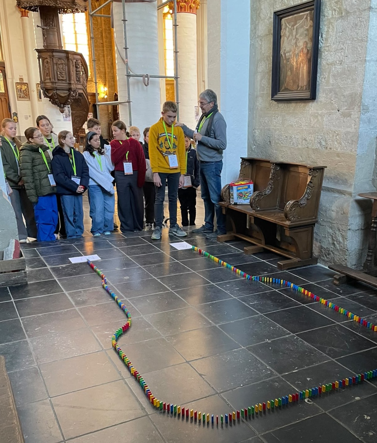 Samen op weg naar het vormsel Een groep jongeren (waarschijnlijk vormelingen of communicanten) samen met een begeleider in een kerk, waarbij een dominoparcours of vloeropdracht wordt gelegd op de stenen vloer.