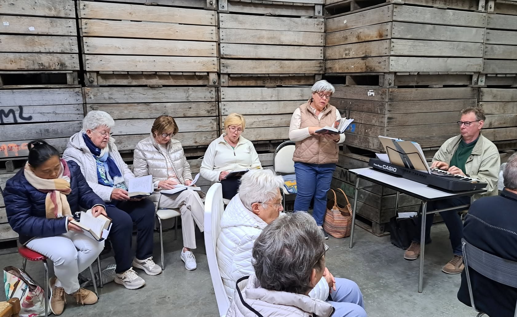 Vruchten der aarde viering Een groep mensen die samen zingt of muziek maakt in een eenvoudige ruimte, vermoedelijk een repetitie of bijeenkomst van een koor.