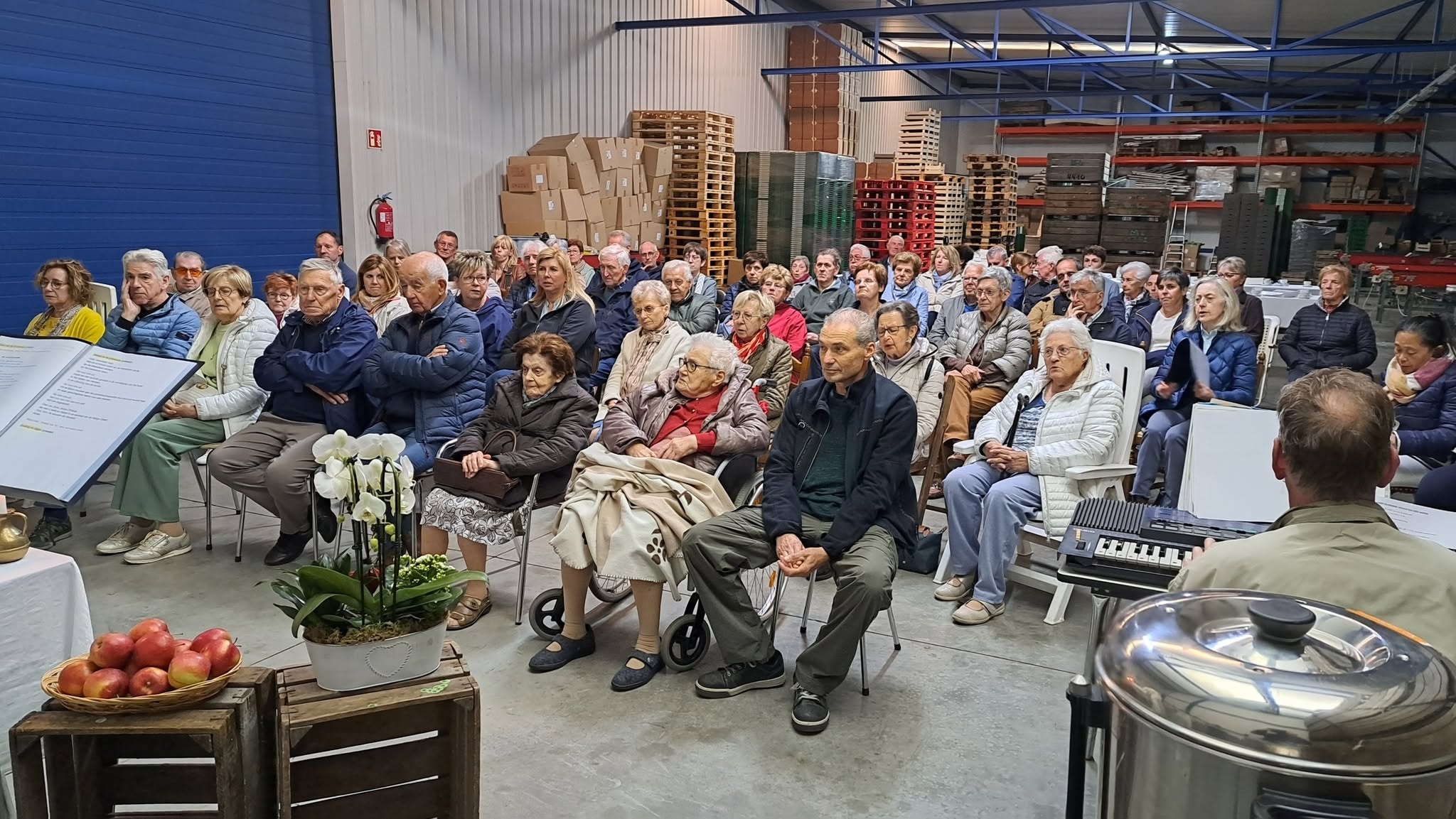 Vruchten der aarde viering Een groep mensen die bijeen is voor een landelijke of lokale viering (zoals een eucharistie- of dankviering voor de "vruchten der aarde") in een loods of schuur.