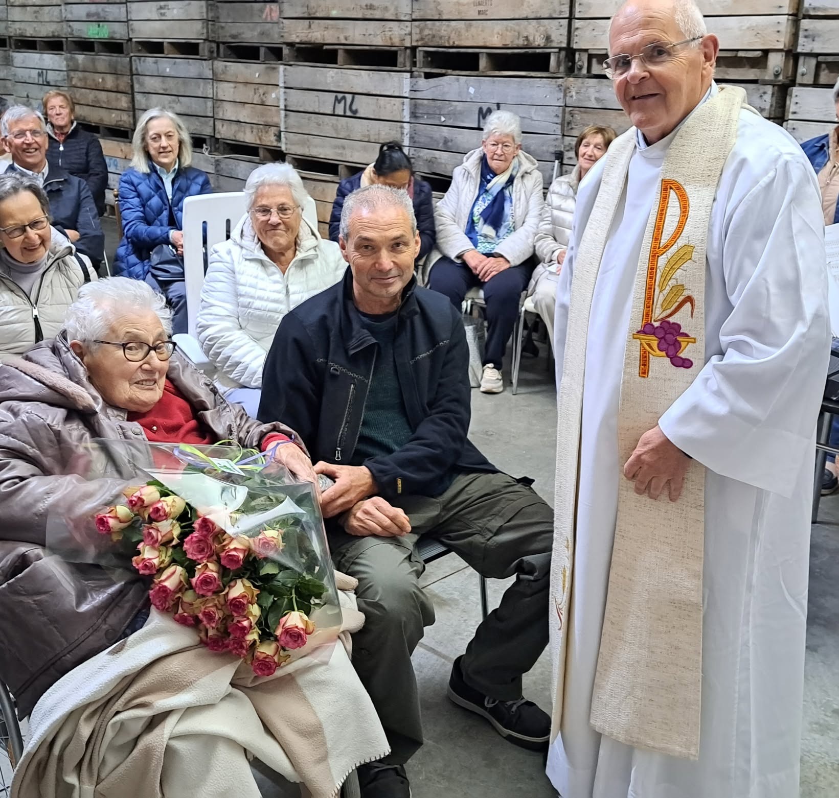 Vruchten der aarde viering Een feestelijke bijeenkomst of huldiging waarbij een oudere vrouw in een rolstoel een groot boeket rozen ontvangt, geflankeerd door een priester in een albe met stola en verschillende toeschouwers die op stoelen zitten voor een wand van houten kisten.