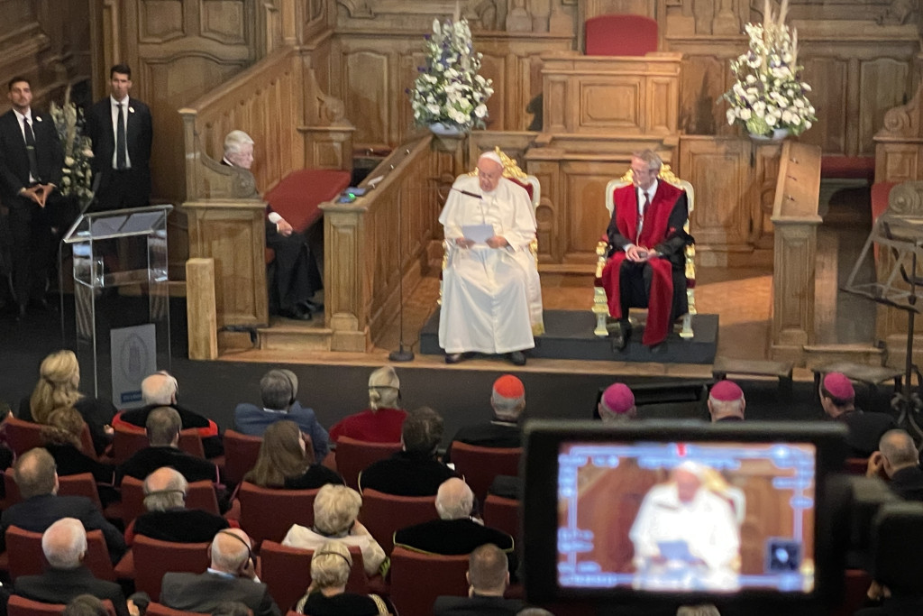 Paus Franciscus aan het woord tijdens de academische zitting in de Promotiezaal van de KU Leuven. Paus Franciscus en rector Luc Sels zitten vooraan in de Promotiezaal van de KU Leuven. Op de voorgrond een camera met hetzelfde beeld.