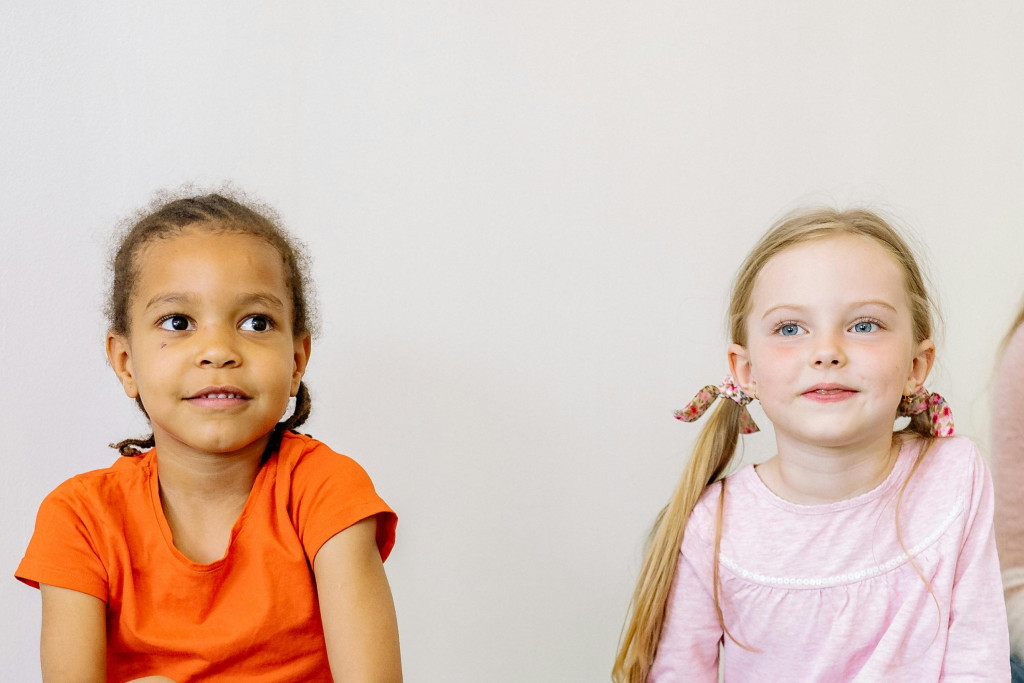 Twee kleutermeisjes zitten met gekruiste benen naast elkaar en kijken vrolijk voor zich uit.