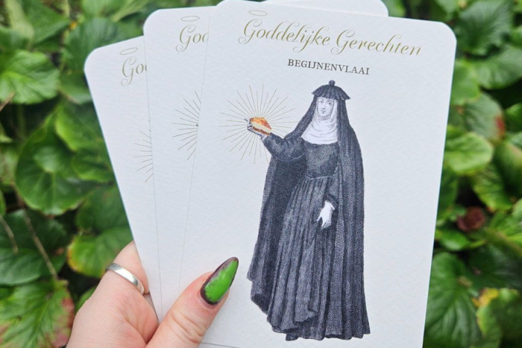 vrouwenhand met opvallend gelakte nagels houdt postkaarten in de hand waarop een begijn met een stuk taart staat afgebeeld.