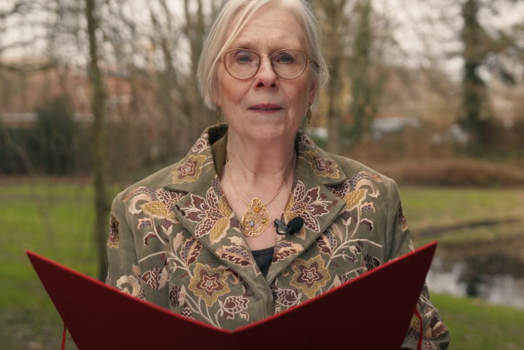 Kolet Janssen leest haar versie van het Zonnelied voor. Kolet Janssen met rode kaft in een park, leest voor.