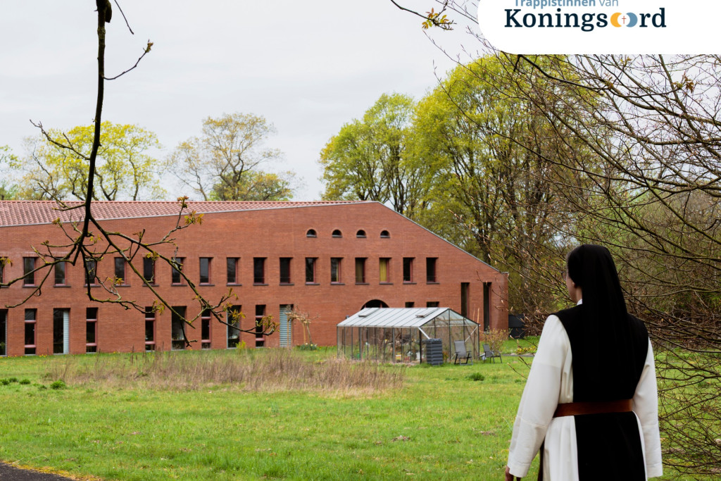 Een zuster vanaf de rug gezien (zwarte kap, witte tuniek, zwart scapulier) stapt richting klooster, een modern, laag gebouw.