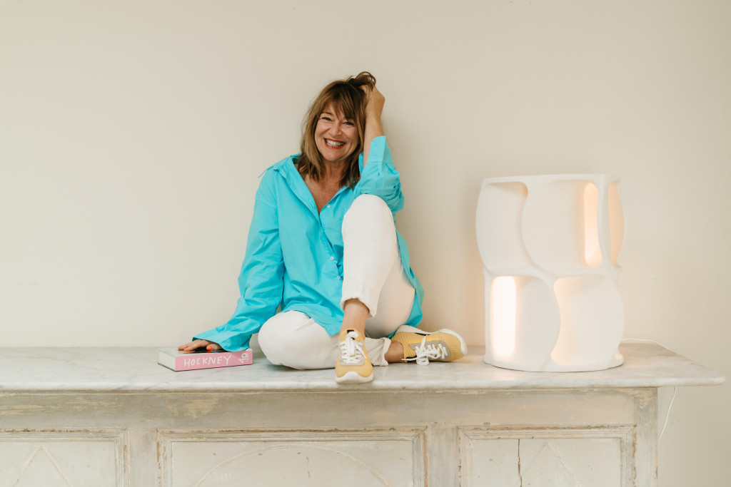 Bie Baert, gulle lach, zit cosy met één been opgetrokken op een antieke kast met designlamp naast haar. Fel appelblauwzeegroen hemd op witte achtergrond.