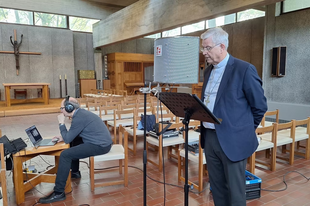 Bisschop Bonny met koptelefoon en audioscherm voor zich. Hij spreekt de teksten in van de podcast, in de Heilige Damiaankerk.