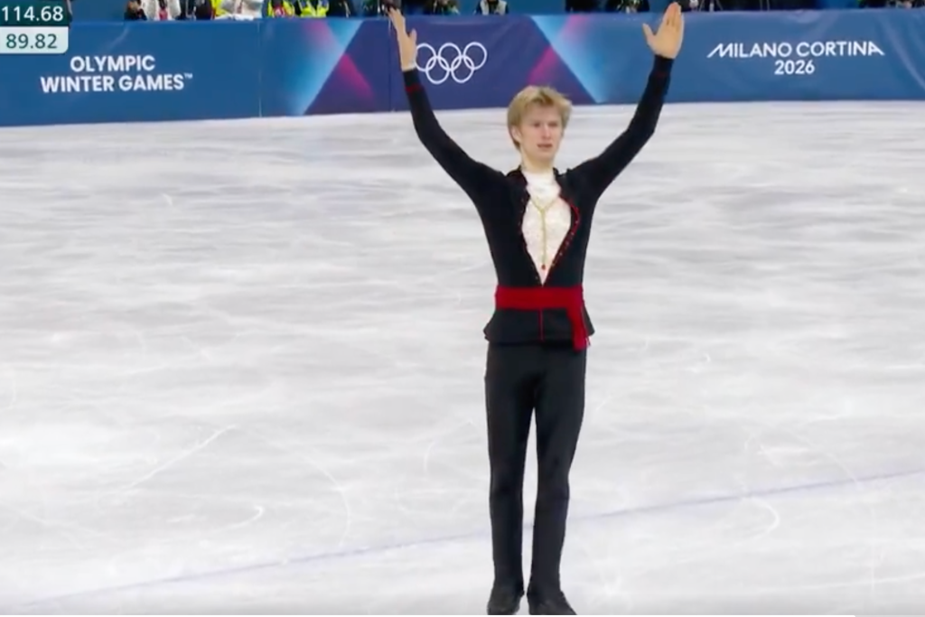 Daniel Grassle groet het publiek, armen in de lucht, op het einde van zijn vrije kür op de Olympische Spelen in Milaan. Onder zijn zwarte top komt een schijnbaar gouden ketting op een witte onderlaag tevoorschijn. 