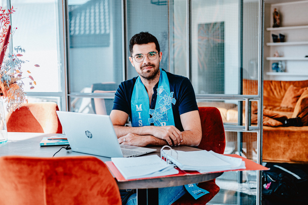 Imran Nawaz aan de computer. Hij heeft een blauwe sjaal met 'Mister Gay Belgium' over de schouders hangen.