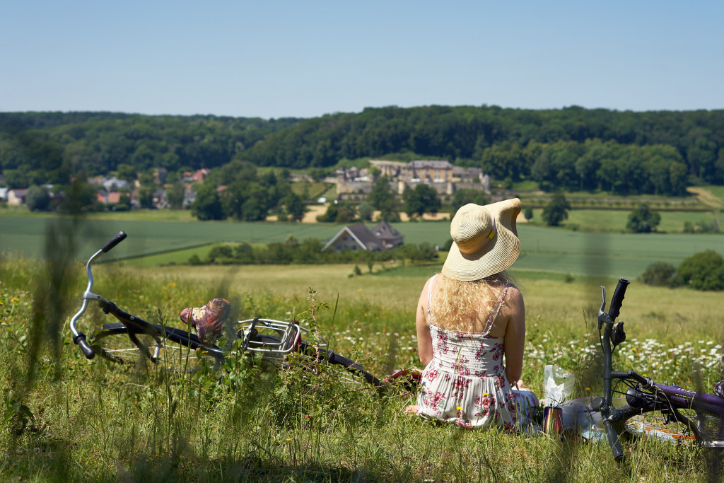 Vrouw met zonnehoed zit in landschap naast neergelegde fiets.