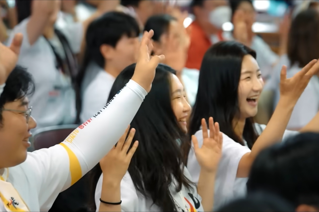 Beeld uit de clip met de hymne van WJD 2027 Seoul Koreanen in wit T-shirt met het logo van WJD 2027 en armen in de lucht, zingend