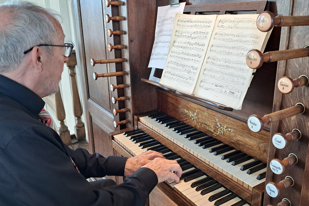 Hugo Adams die aan de speeltafel van een pijporgel zit en op twee manualen (klavieren) speelt, te midden van bladmuziek en herkenbare registertrekkers aan de zijkant.