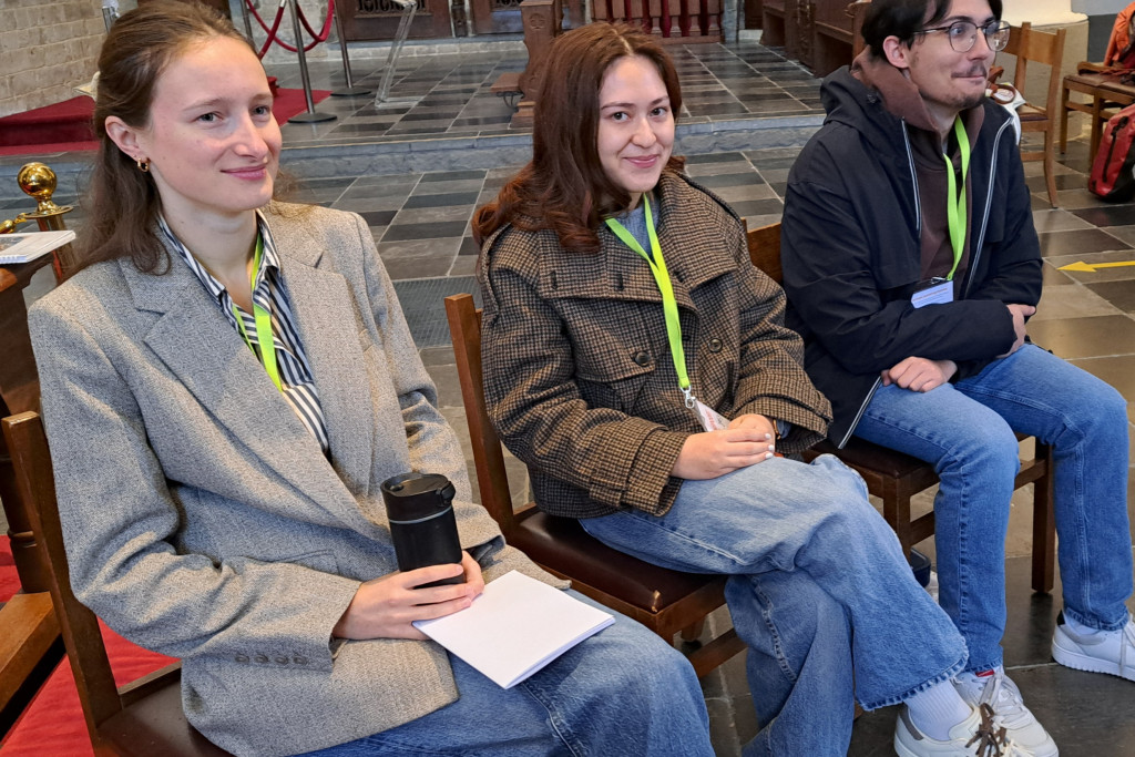 Drie jonge mensen die in een kerkgebouw of een soortgelijke zaal op houten stoelen zitten en een lanyards/badges dragen,